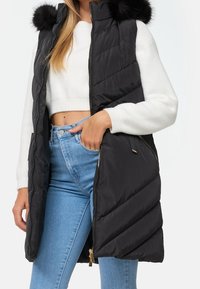 Veste sans manches noire matelassée avec fermeture éclair et capuche bordée de fourrure, présentant un matelassage en forme de chevrons et des poches latérales, portée avec un jean bleu clair.