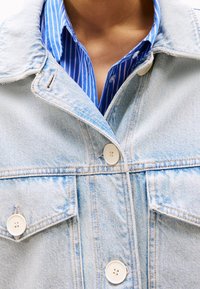 Light blue denim jacket with a collar, two front pockets, and white buttons, layered over a blue and white striped shirt.