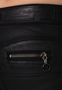 Jeans negros con un pequeño bolsillo con cremallera y herrajes metálicos, un parche de cuero con el nombre de la marca y detalles de costura sutiles.