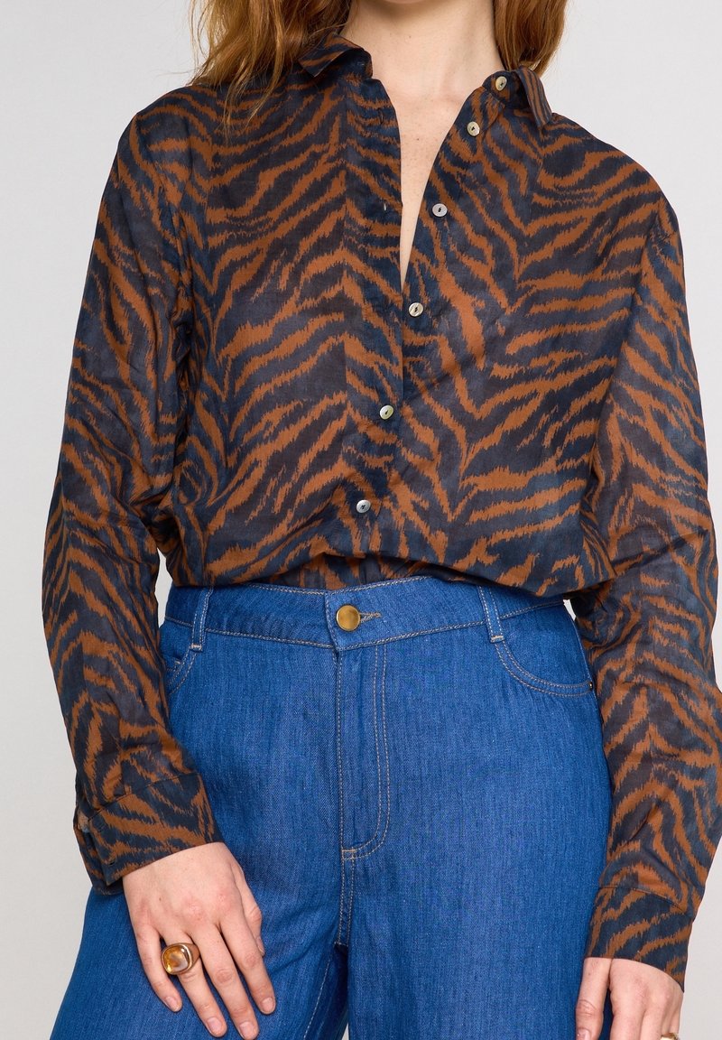 Blouse boutonnée à rayures tigre marron et noire associée à un jean bleu taille haute, bague en ambre visible à la main gauche.