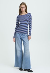 Stribet langærmet top i blå og mørkeblå, parret med bredbenede lyseblå jeans. Modellen står imod en neutral baggrund.