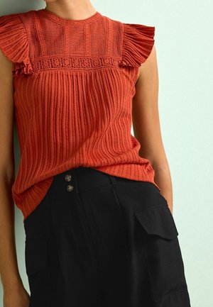 Kobieta nosząca rdzawo-pomarańczową, teksturowaną, bezrękawną bluzkę z plisowanymi detalami na ramionach oraz czarne spodnie z wysokim stanem z kieszeniami na guziki i klapy.
