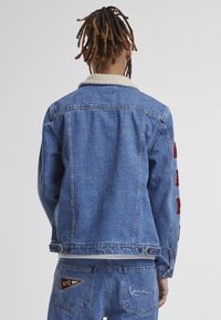 Blue denim jacket with a white shearling collar, red embroidered letters on sleeves, and a straight cut. Worn with matching blue jeans.