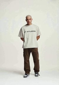 Ung man med kort blont hår som bär en vit t-shirt med texten "2Y STUDIOS", bruna cargo byxor och svarta och vita sneakers, stående med händerna bakom ryggen.