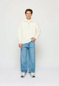 Jauns vīrietis valkā baltu puszipa flīsa džemperi, brīvu zilo biksu un baltas kedas, stāvot uz viendabīga gaiša fona.