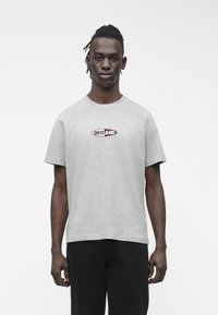 Pilkas trumpomis rankovėmis marškinėlis su juoda ir raudona „Guess Jeans“ logotipu ovalo formos dizaine krūtinės srityje. Minkšta medvilnė.