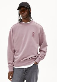 Sweatshirt lavande avec un col rond et des poignets côtelés, arborant un logo texturé sur la poitrine. Associé à un jean gris.