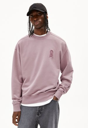 Sweatshirt lavande avec un col rond et des poignets côtelés, arborant un logo texturé sur la poitrine. Associé à un jean gris.