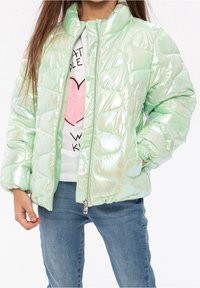 Enfant portant une veste matelassée vert clair brillante, un jean et une chemise blanche avec un cœur rose et un texte noir, mains dans les poches.