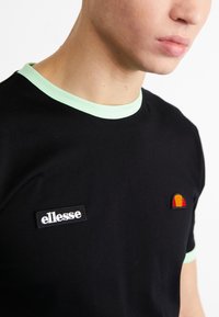 Black t-shirt with a light green ribbed collar, featuring an embroidered logo and a colorful accent on the chest. Smooth fabric texture.