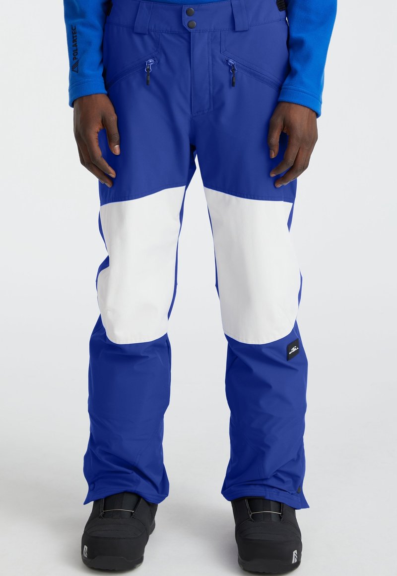 Person wearing blue and white snow pants with zippered pockets and black winter boots, standing against a plain background.