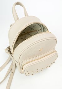 Zaino in pelle beige con forma arrotondata, due scomparti con cerniera, hardware dorato e accenti di borchie nella parte anteriore inferiore.