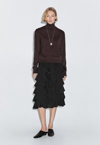 Brown turtleneck sweater paired with a black ruffled midi skirt. The skirt features multiple layers, and the outfit is completed with black slip-on shoes.
