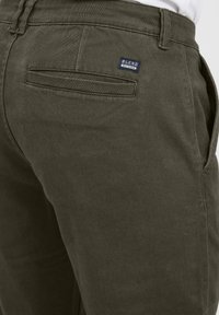 Olive green pants made from fabric with a smooth texture. Features a small brand label on the back pocket and standard side pockets.