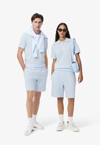 Polo shirt e pantaloni corti di colore azzurro, realizzati in un tessuto morbido e con una vestibilità rilassata. Gli accessori includono una borsa azzurra e occhiali da sole.