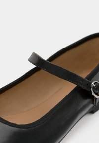 Ballerina in pelle nera con punta arrotondata, caratterizzata da un sottile cinturino e fibbia color argento. Texture liscia e lucida con interno beige chiaro.