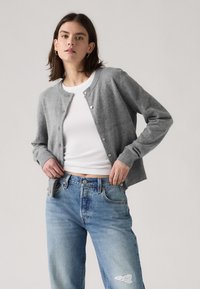 Frau trägt grauen, geknöpften Cardigan über weißem, geripptem Top und blauen, ausgewaschenen Jeans, steht vor hellgrauem Hintergrund.