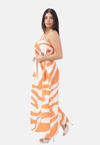 Donna in un maxi dress senza maniche, arancione e bianco con un motivo, che tiene la cintura, indossando sandali bianchi con tacco alto.