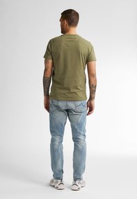 T-shirt en coton vert olive avec un col rond, associé à un jean en denim bleu clair. Les tatouages du modèle sont visibles sur les bras.