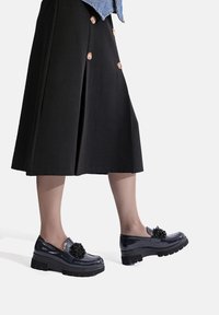 Scarpe slip-on in pelle verniciata nera con suola spessa, caratterizzate da un pom-pom decorativo nero nella zona della punta. Texture liscia, forma arrotondata.