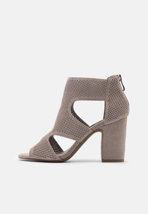 Åbne højhælede sandaler i lysegrå ruskind med udskårne mønstre og lynlås bagpå. Kraftig hældesign.