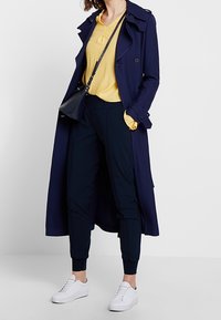 Femme portant un long trench-coat bleu marine, un blouse jaune, un pantalon taillé bleu marine, des baskets blanches et portant un sac bandoulière noir.