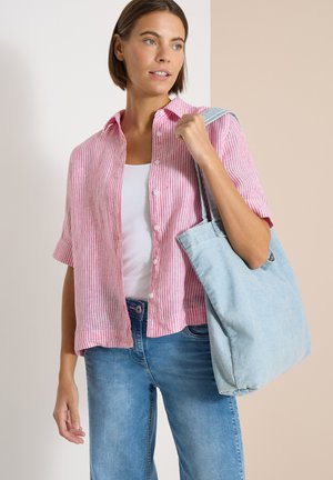 Mujer con camisa rosa a rayas abotonada y vaqueros azules, llevando un gran bolso tote azul claro colgado del hombro.