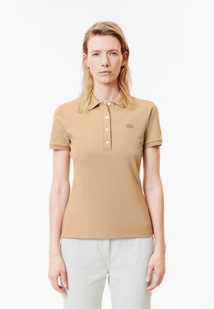 Polo shirt beige con maniche corte, colletto a tre bottoni e logo discreto sul petto. Abbinato a pantaloni chiari. Texture liscia.
