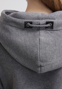 Harmaa huppari, jossa on teksturoitu kangas, kiristysnauha ja mustat silmukat säädettävää istuvuutta varten.