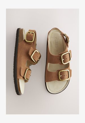 Sandales en daim marron avec trois brides ajustables munies de boucles dorées, une semelle intérieure légère et rembourrée, et une semelle en caoutchouc noir.