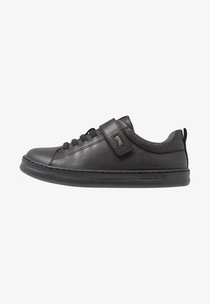 Black leather sneaker with rounded toe, lace-up front, and adjustable Velcro strap. Features a rubber sole and stitched accents.