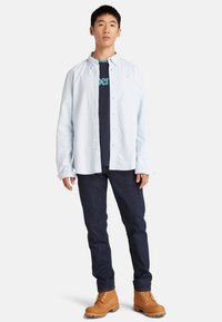 Lichtblauw overhemd met knopen over een marineblauw T-shirt, donkere jeans en tan laarzen tot de enkel. Het overhemd heeft een relaxte pasvorm en lange mouwen.