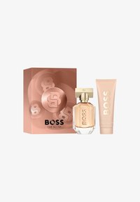 El set de regalo BOSS The Scent incluye una botella de vidrio transparente con una tapa dorada y un tubo rosa, ambos con el logotipo de BOSS, en una caja de color rosa dorado.