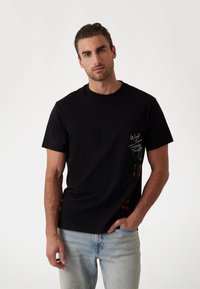 Sort bomulds t-shirt med rund halsudskæring og korte ærmer, med hvid tekst på forsiden og farverigt grafisk design på siden.