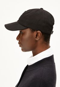 Schwarze Kappe aus Baumwolle mit einem gebogenen Schirm, sechs-Panel-Design und gesticktem Logo. Das Modell trägt einen dunklen Pullover und ein Kragenhemd.
