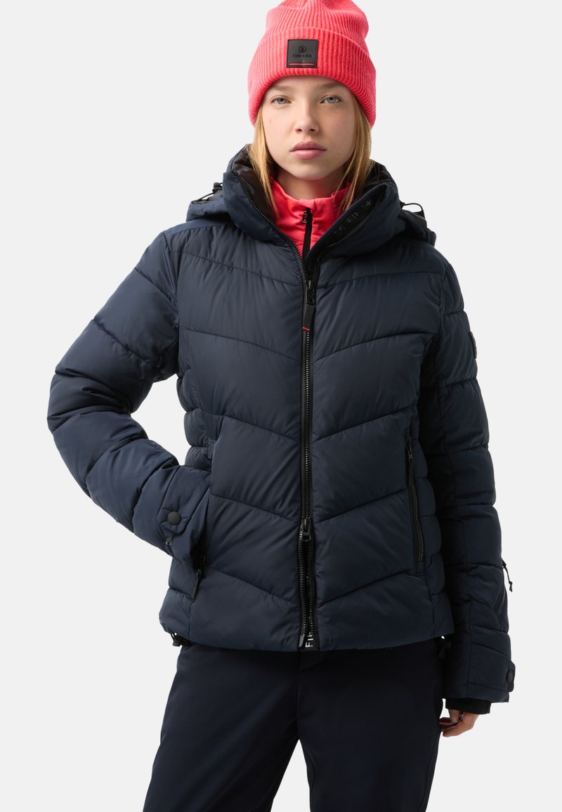 Marineblaues Steppjacke aus isolierendem Material, horizontale Steppung, mit Reißverschlusstaschen und abnehmbarer Kapuze. Kombiniert mit einer pinken Strickmütze.