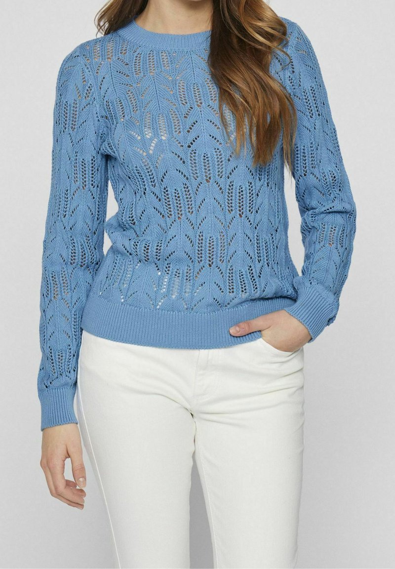 Light blue knitted sweater with a lace pattern, ribbed neckline and cuffs, featuring long sleeves and a fitted waist. Paired with white pants.