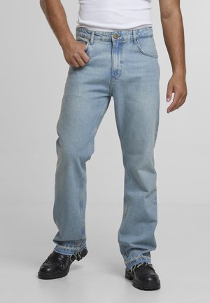 Mann trägt hellblaue Jeans mit geradem Bein und ausgefransten Säumen, schwarze klobige Schuhe und ein weißes ärmelloses Oberteil, steht vor einem grauen Hintergrund.