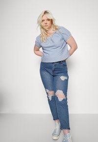 Blusa de mangas curtas azul e branca com riscas, com decote ajustado, combinada com jeans azuis com desgaste e rasgos significativos e ténis azuis.