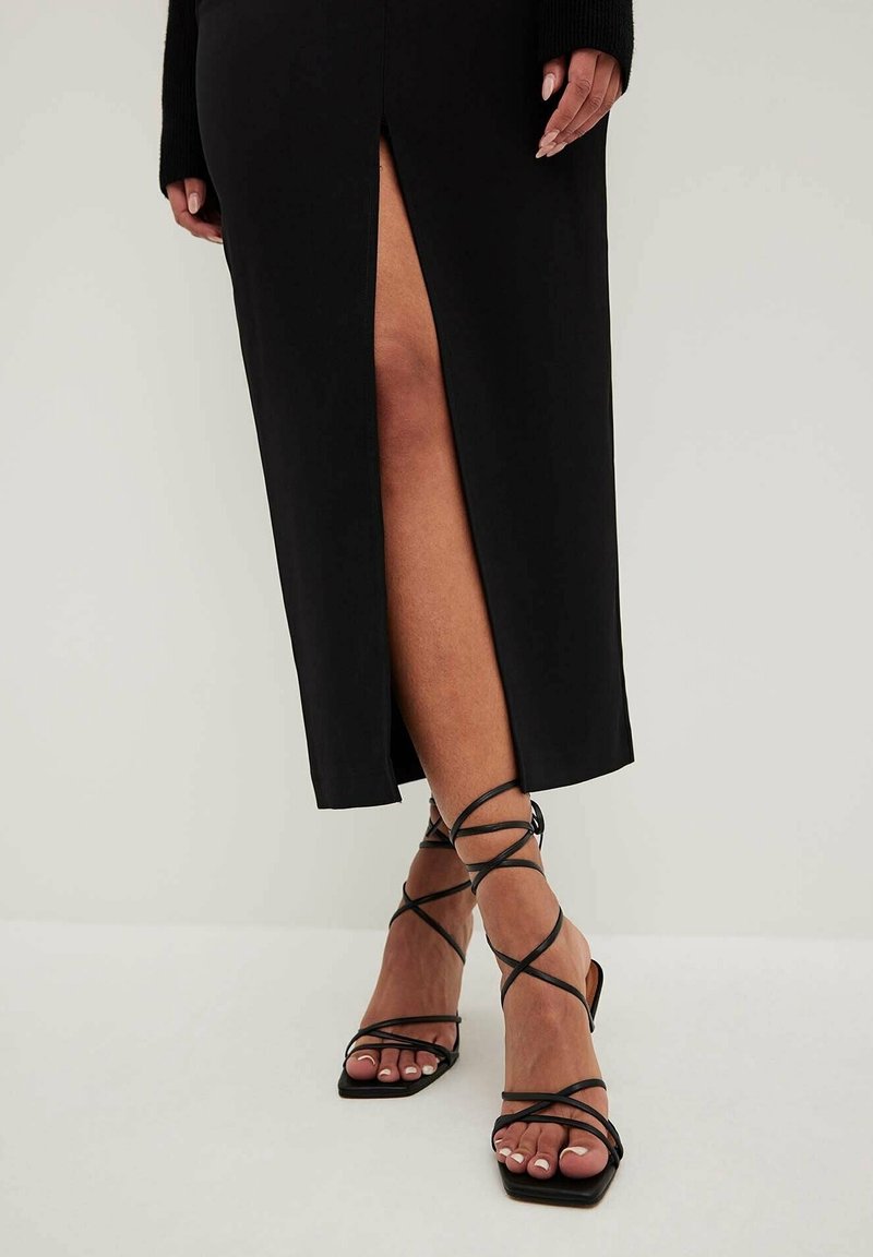 Femme portant une longue jupe noire avec une fente à l'avant et des sandales à talons hauts noires à lanières, montrant des jambes croisées sur un sol blanc.