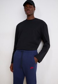 Sort langærmet T-shirt parret med navy sweatpants med et rødt logo, snoreliv og lommeaccenter.