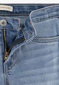 Blugi super skinny bleu deschis din denim, prevăzuți cu nasture metalic și fermoar, cu cusături vizibile și textură netedă.