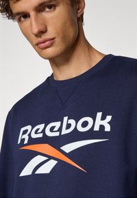 Námornícky sveter s rebrovaným okrajom okolo krku, s veľkým bielym logom Reebok a oranžovým akcentovým grafikou. Hladká, štruktúrovaná tkanina.