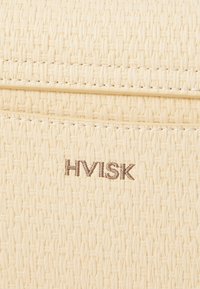 Bež tkanu torbu s teksturiranom površinom, koja ima šivene detalje i utisnuti "HVISK" logo u smeškastoj boji sprijeda.