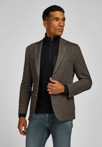 Brown houndstooth blazer with notched lapels, two gold button closures, and a navy high collar sweater underneath, paired with denim jeans.