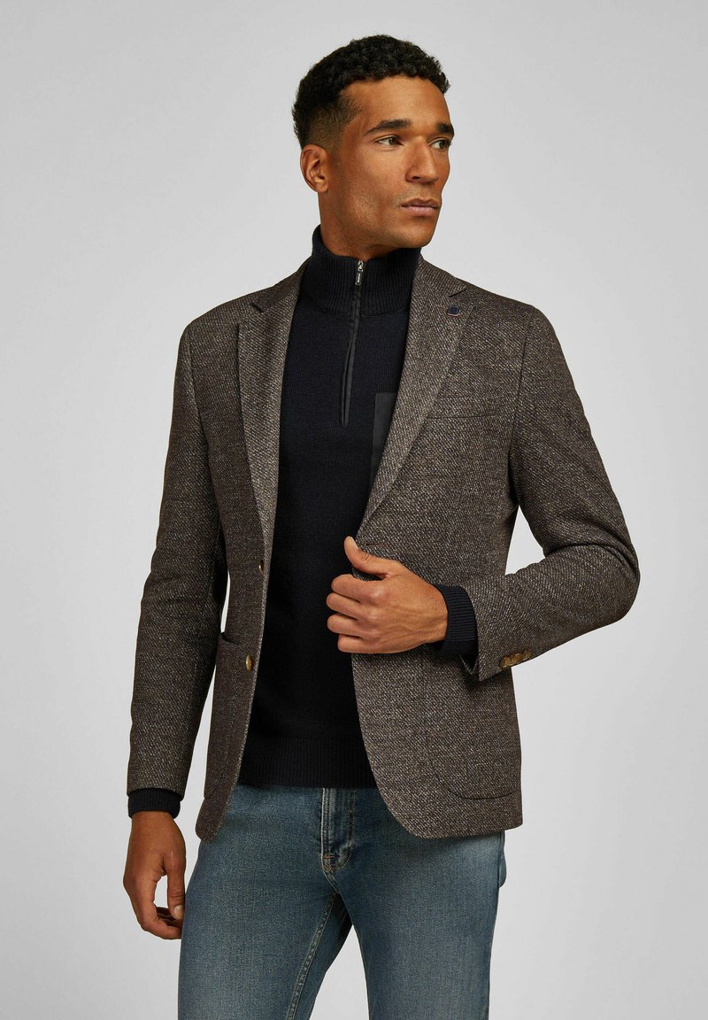 Brown houndstooth blazer with notched lapels, two gold button closures, and a navy high collar sweater underneath, paired with denim jeans.
