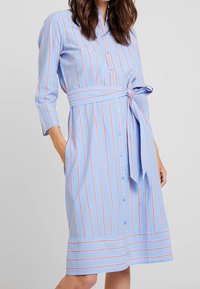 Niebieska sukienka w paski z czerwonymi paskami, z guzikami z przodu, rękawy do łokci, związywana w talii, z kieszeniami i długością do kolan.