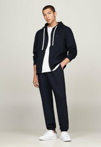 Marineblauer Baumwollhoodie mit weißen Kordeln undFronttaschen, kombiniert mit passenden Jogginghosen und weißen Sneakers. Schlichtes, sauberes Design.