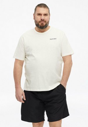 Mężczyzna z brodą i związanymi do tyłu włosami, ubrany w białą koszulkę "Après Surf" i czarne szorty, stoi z jedną ręką w kieszeni.