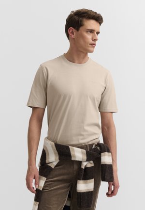Mann trägt beige Kurzarmhemd und braunen gestreiften Pullover um die Taille gebunden, schaut seitlich vor einfarbigem Hintergrund.